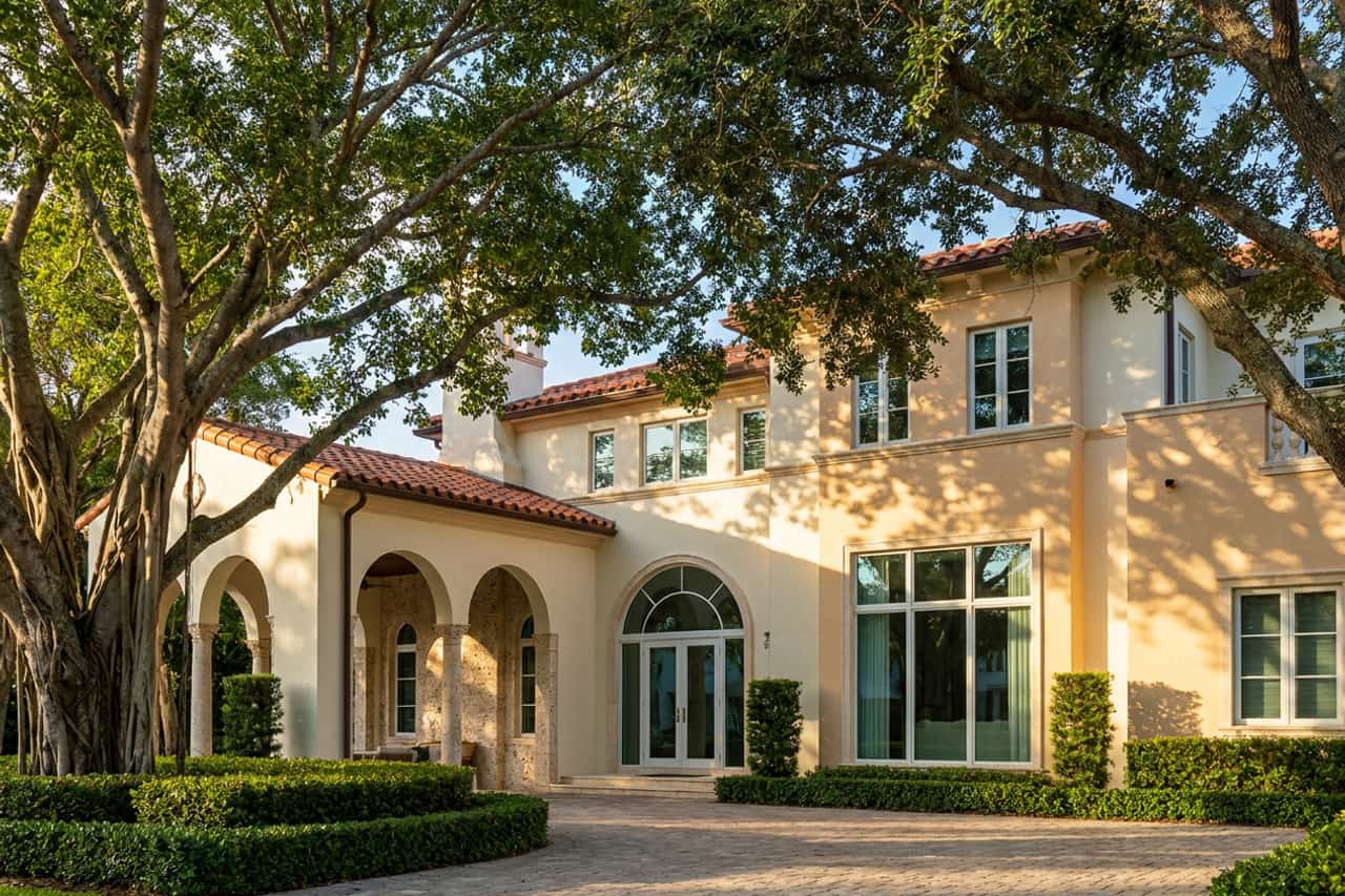 A Mediterranean-revival residence with a coral-rock accent wall under a banyan canopy, in the City Beautiful style of Coral Gables, with impact-rated windows and an arched glass door